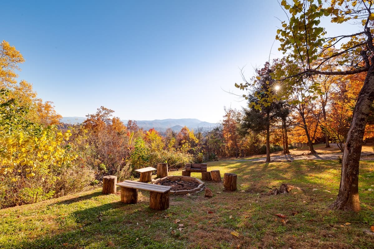 Fire pit area with log bench and stump seating surrounded by golden fall foliage with mountain views and sunburst
