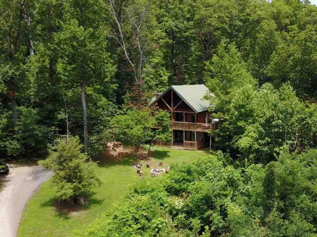 Aerial view of two-story log cabin with green roof on one acre lot surrounded by woods with fire pit and gravel driveway